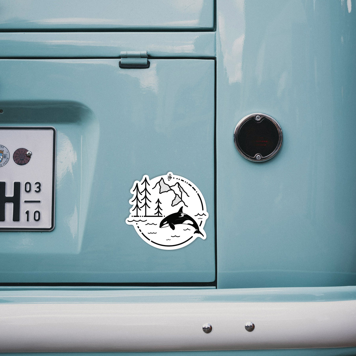 4 inch vinyl decal of a black line art circle connecting pine trees, mountains, a bee and an orca leaping from ocean waves on the back of a van.