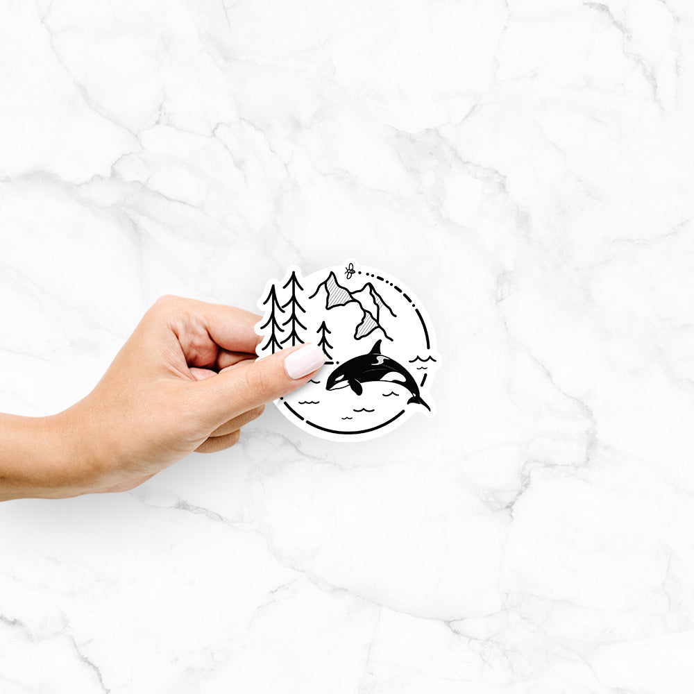 Hand holding a 4 inch vinyl decal of a black line art circle connecting pine trees, mountains, a bee and an orca leaping from ocean waves.