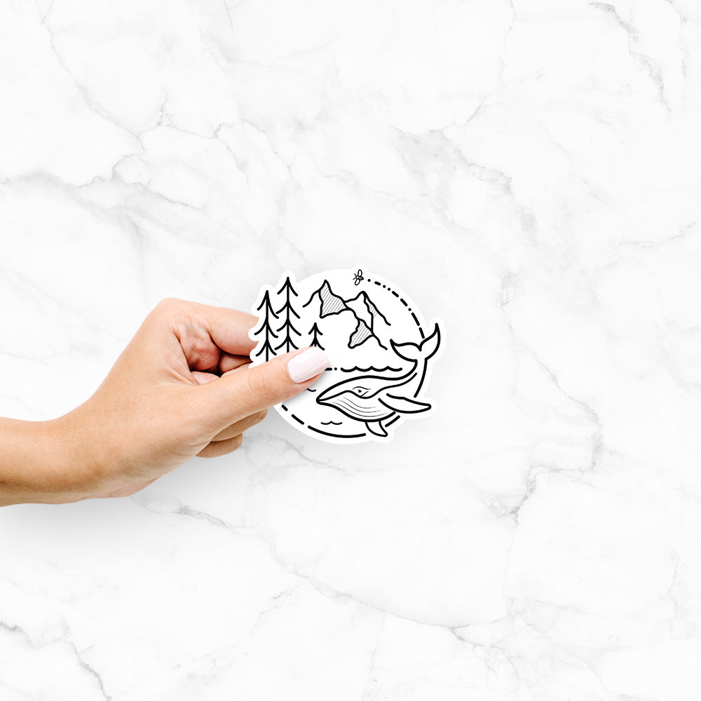Hand holding a 4 inch vinyl decal of a black line art circle connecting a mountain range, pine trees, a bee and a whale swimming in ocean waves.