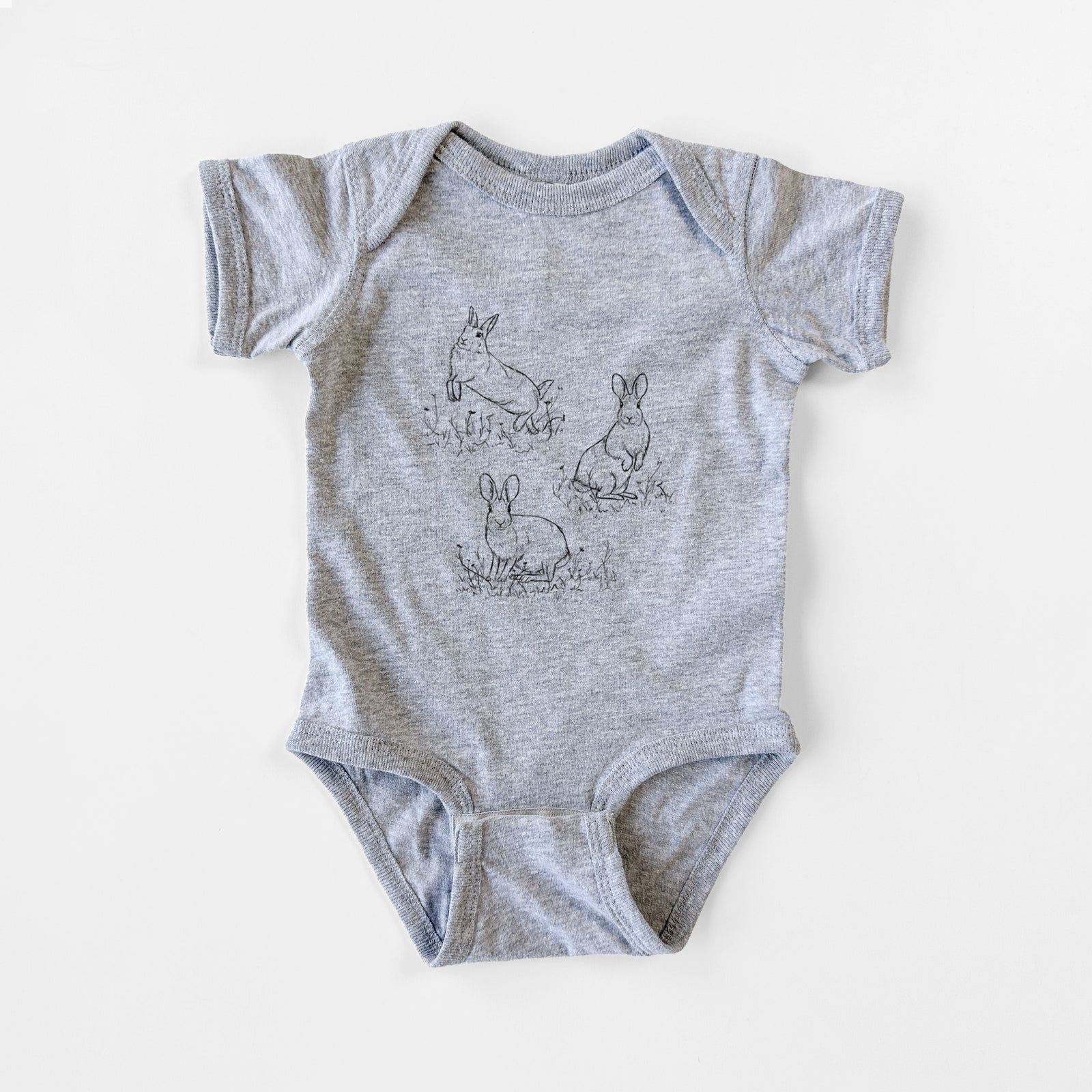 Grey baby bodysuit laid flat, printed with a black ink illustration of three cottontail rabbits among grass and flowers.