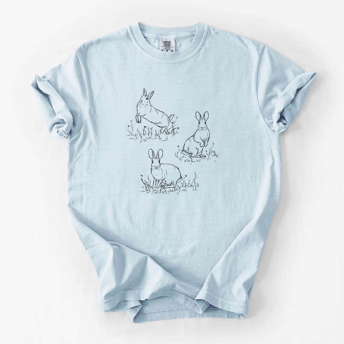 Chambray blue Comfort Colors t-shirt laid flat, printed with a black ink illustration of three cottontail rabbits among grass and flowers.