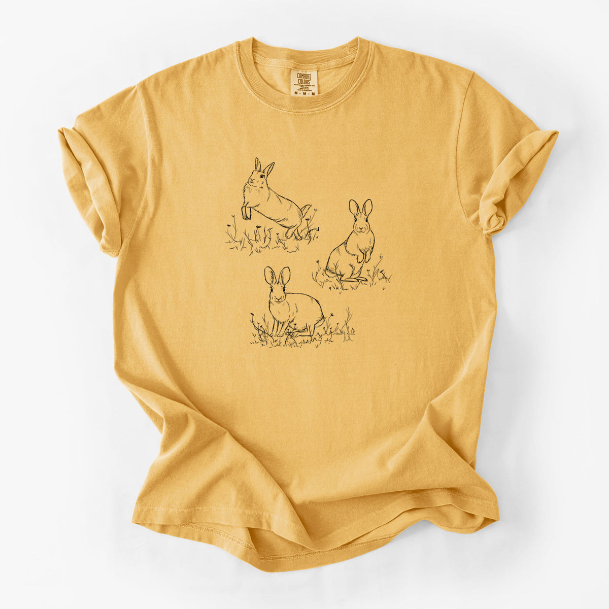 Golden wheat yellow Comfort Colors t-shirt laid flat, printed with a black ink illustration of three cottontail rabbits among grass and flowers.