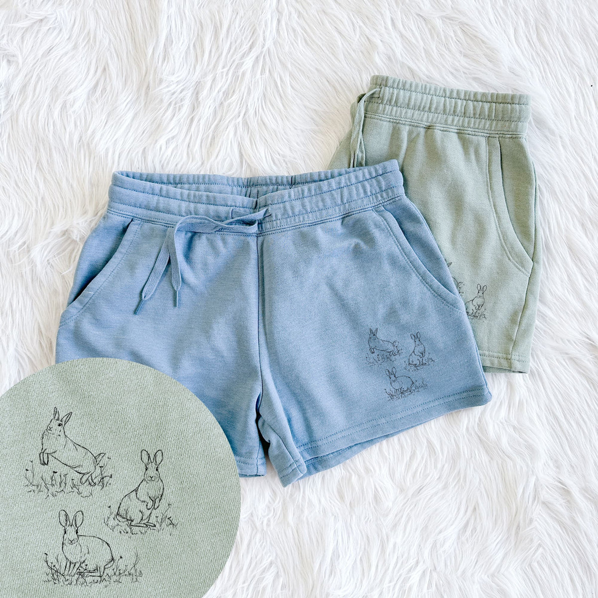 Two pairs of Cali Wave shorts in different colors printed with a black ink illustration of three cottontail rabbits among grass and flowers, with a close-up of the print.