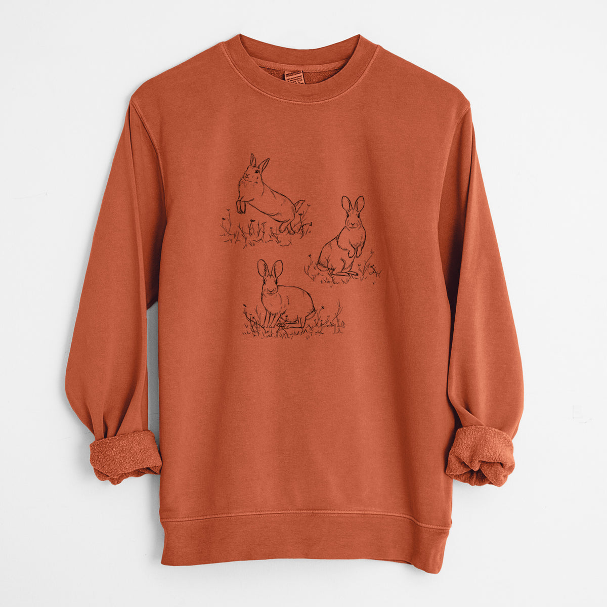 Amber orange pigment dyed crewneck sweatshirt laid flat with the sleeves rolled, printed with a black ink illustration of three cottontail rabbits among grass and flowers.