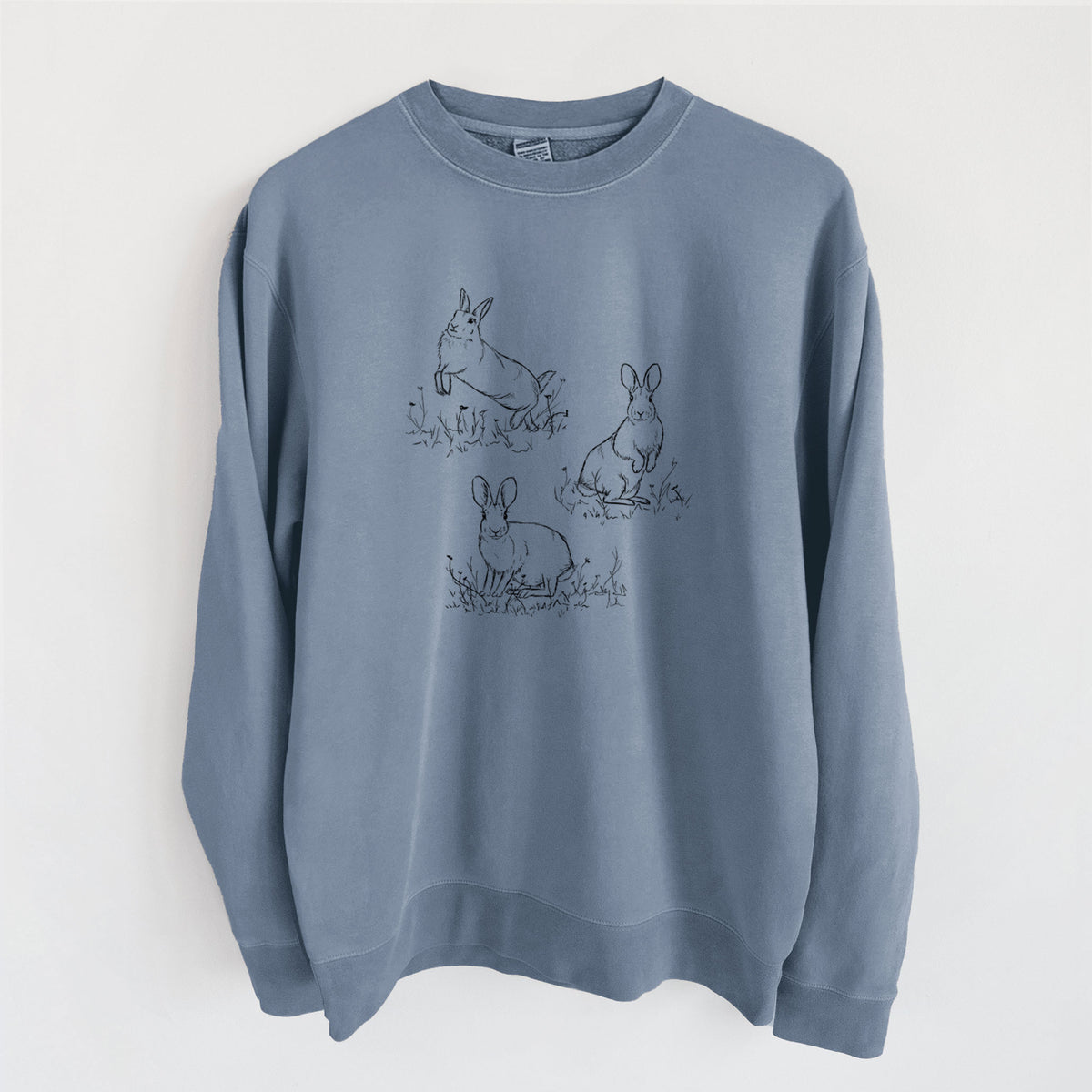 Slate blue pigment dyed crewneck sweatshirt laid flat, printed with a black ink illustration of three cottontail rabbits among grass and flowers.