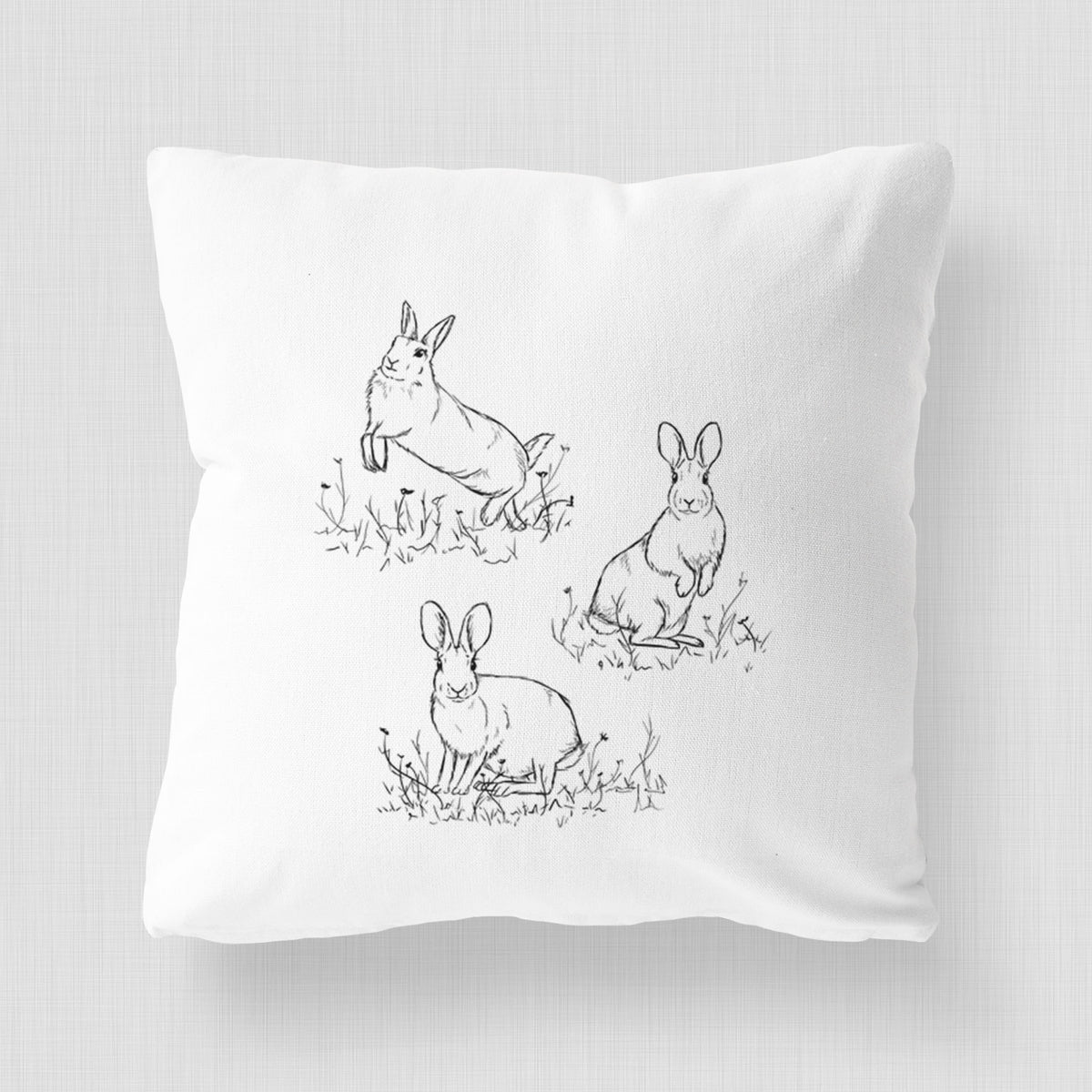 White pillow cover printed with a black ink illustration of three cottontail rabbits among grass and flowers.