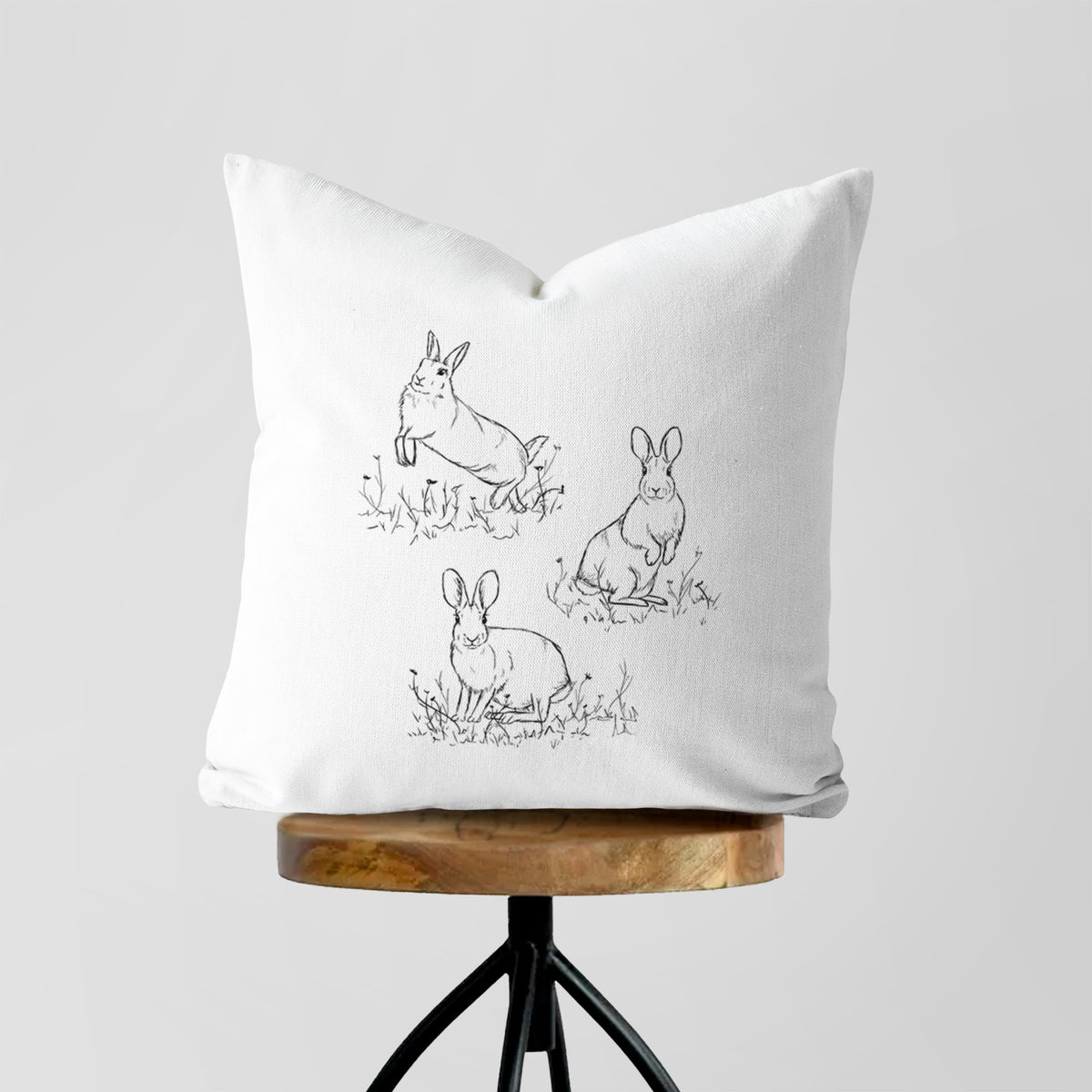 White pillow cover printed with a black ink illustration of three cottontail rabbits among grass and flowers, on a wooden stool.