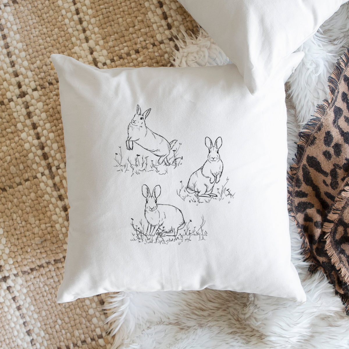 White pillow cover printed with a black ink illustration of three cottontail rabbits among grass and flowers, on a woven chair.