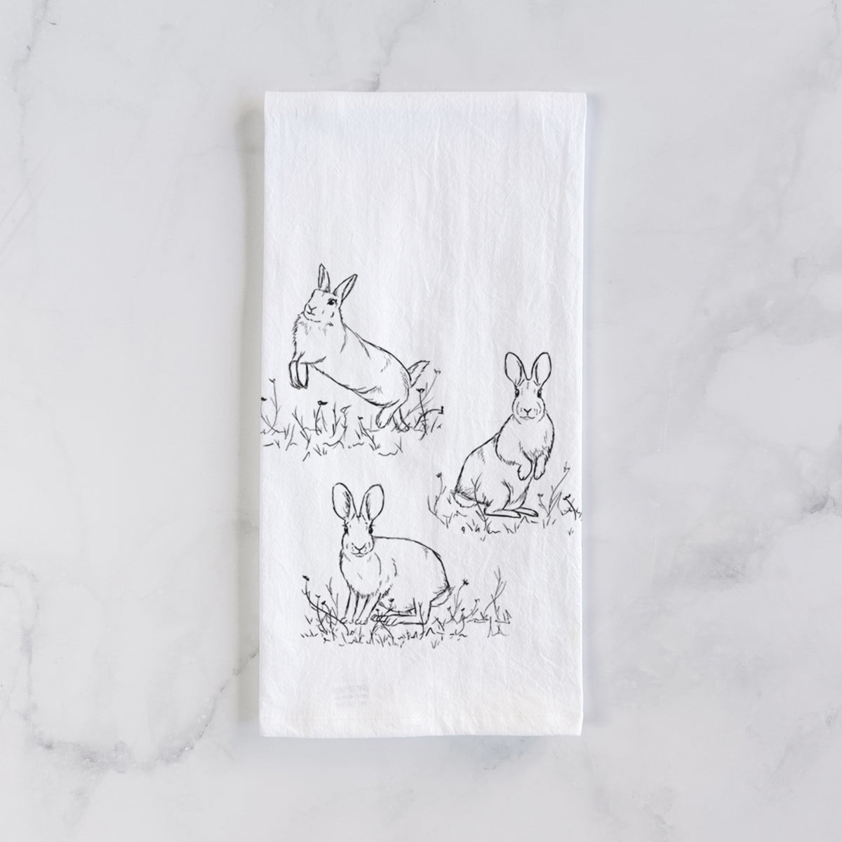 White cotton tea towel laid flat on marble, printed with a black ink illustration of three cottontail rabbits among grass and flowers.