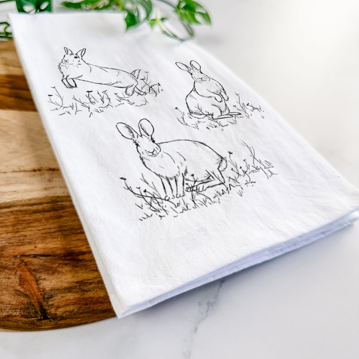 Close-up of a folded white tea towel on a wooden board, showing a black ink illustration of three cottontail rabbits among grass and flowers.