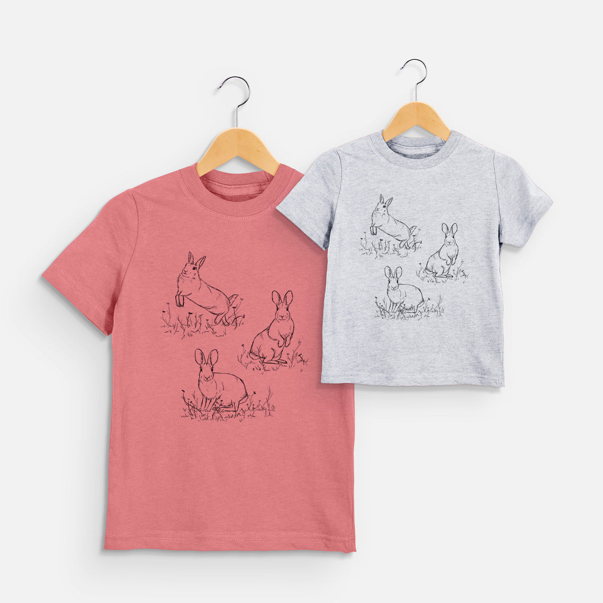 Two youth t-shirts in different colors on hangers, printed with a black ink illustration of three cottontail rabbits among grass and flowers.