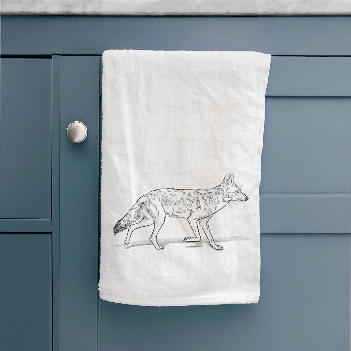 White hand towel printed with a black ink illustration of a coyote trotting, hanging on a cabinet door.