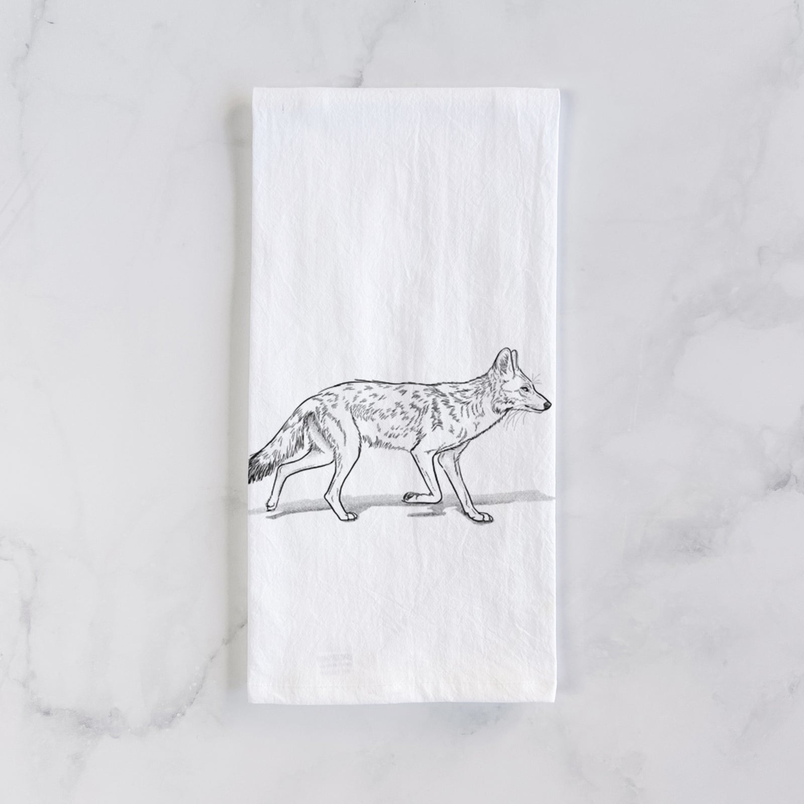 White cotton tea towel printed with a black ink illustration of a coyote trotting, on a marble counter beside a round wooden cutting board and greenery.