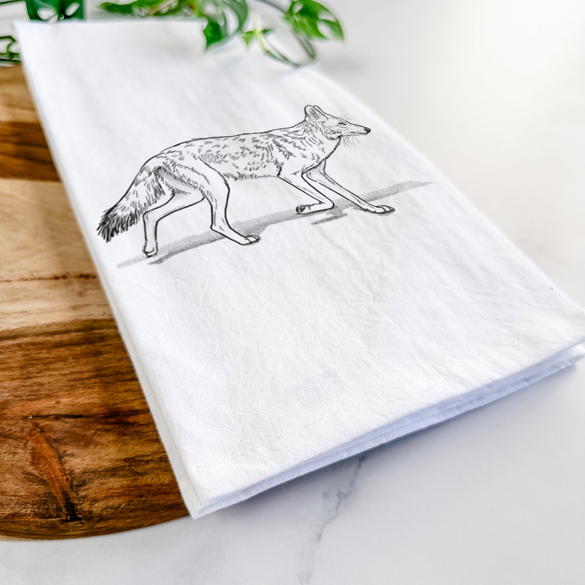 Close-up of a folded white tea towel on a wooden board, showing a black ink illustration of a coyote trotting.