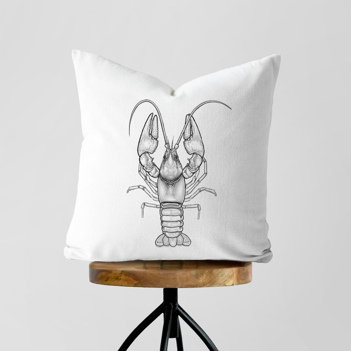 White pillow cover printed with a detailed black ink illustration of a crayfish with its claws raised, seen from above, on a wooden stool.