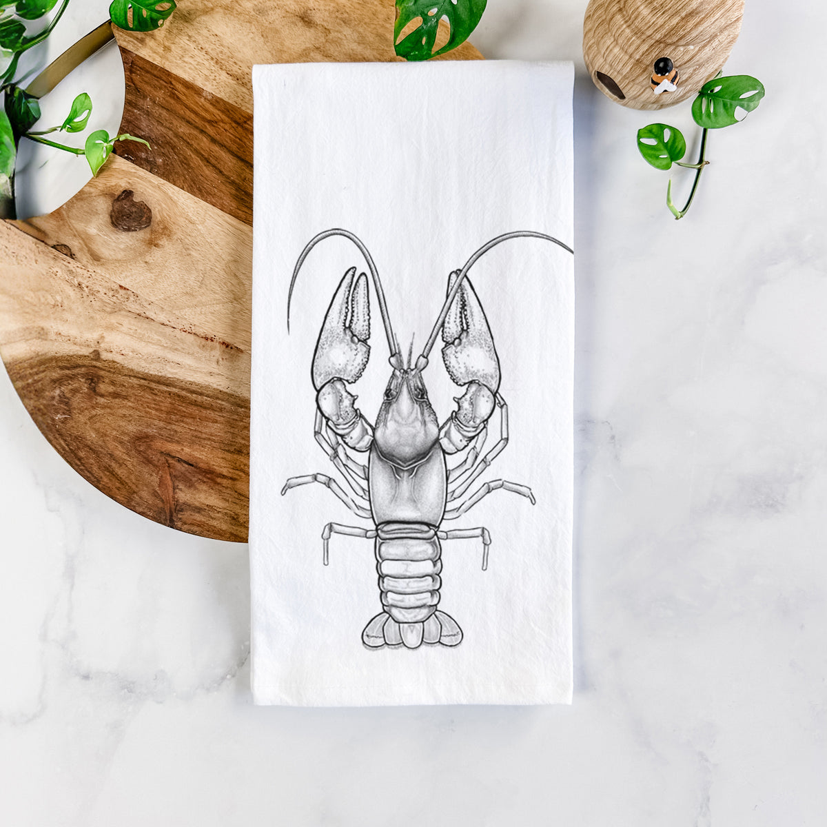 White cotton tea towel printed with a detailed black ink illustration of a crayfish with its claws raised, seen from above, on a marble counter beside a round wooden cutting board and greenery.