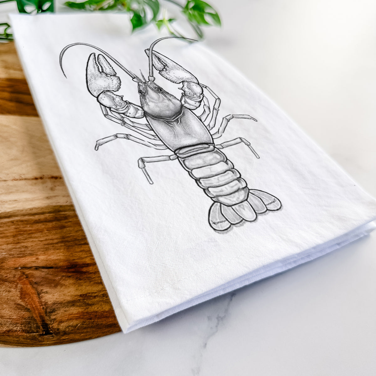 Close-up of a folded white tea towel on a wooden board, showing a detailed black ink illustration of a crayfish with its claws raised, seen from above.