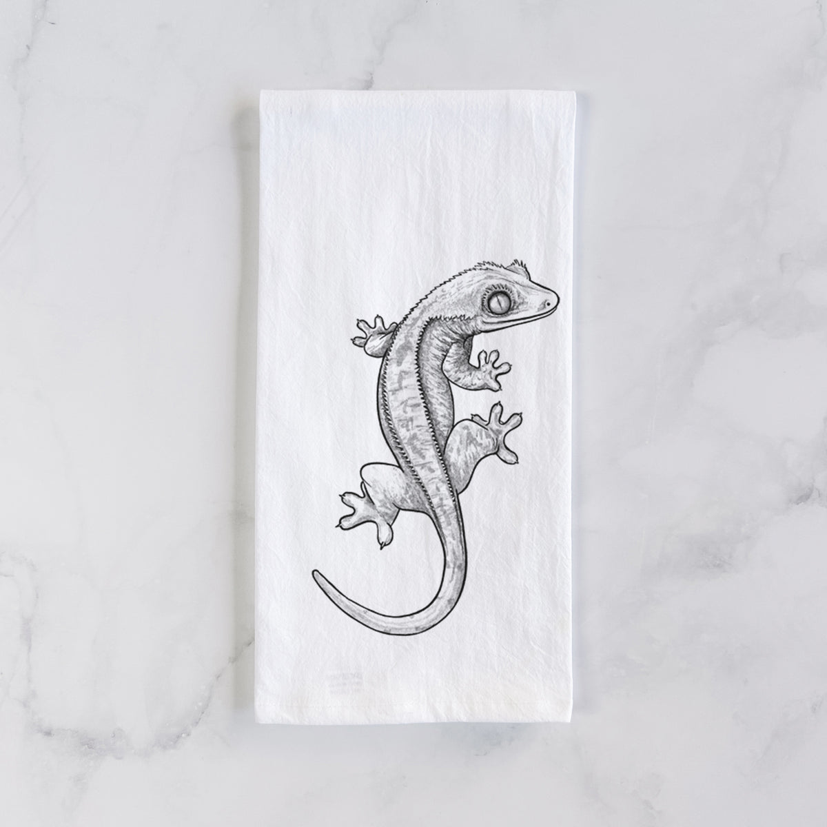 White cotton tea towel laid flat on marble, printed with a detailed black ink illustration of a crested gecko climbing.