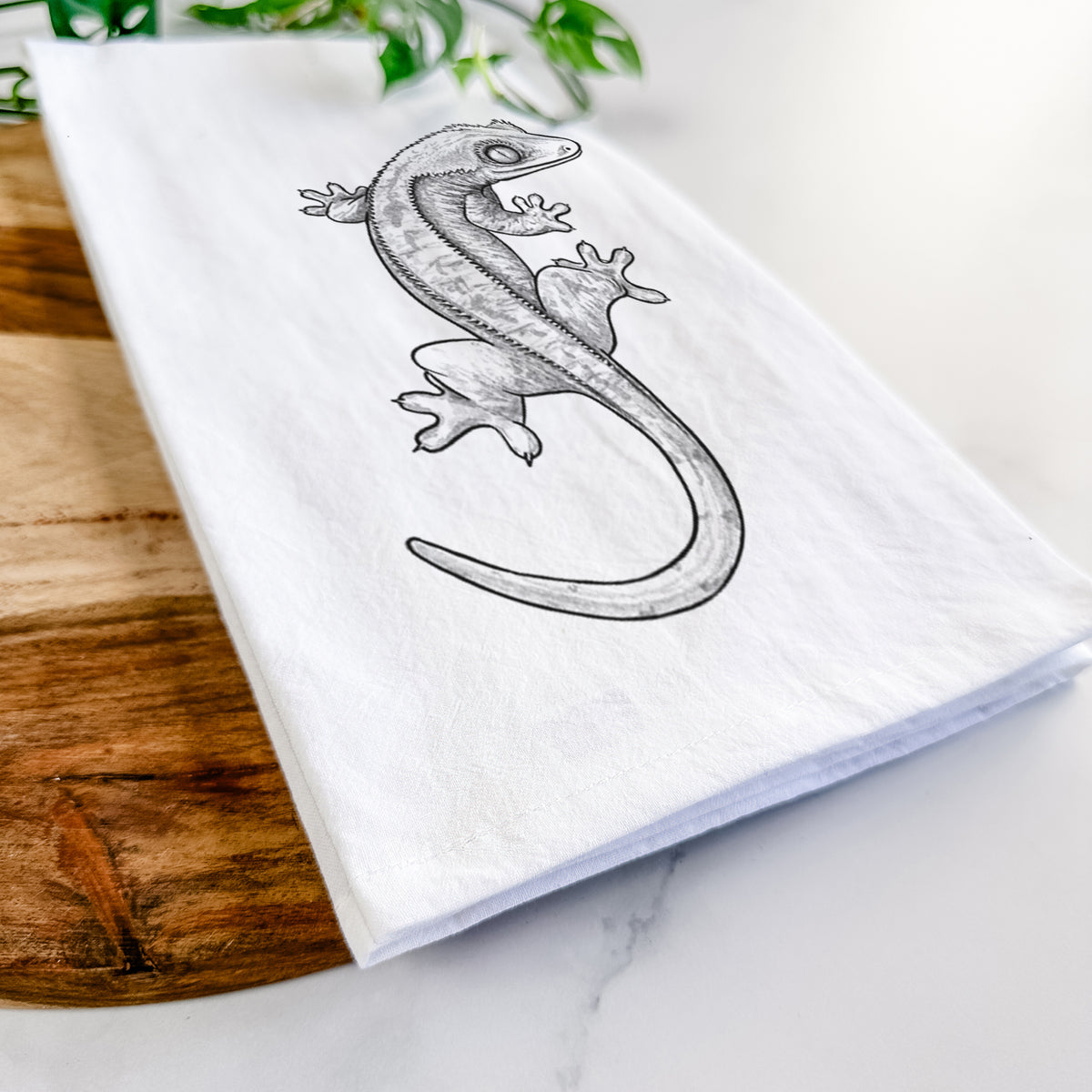 Close-up of a folded white tea towel on a wooden board, showing a detailed black ink illustration of a crested gecko climbing.