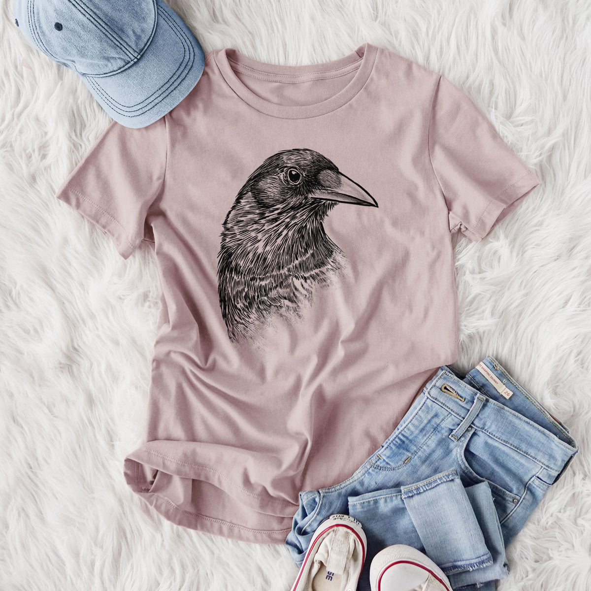 Women's relaxed fit cotton t-shirt printed with a detailed black ink portrait of a crow's head in profile, styled flat on a white faux fur rug with jeans.