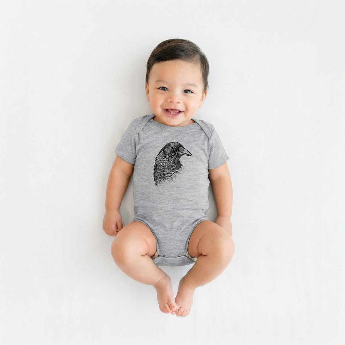 Smiling baby boy wearing a grey baby bodysuit printed with a detailed black ink portrait of a crow's head in profile.