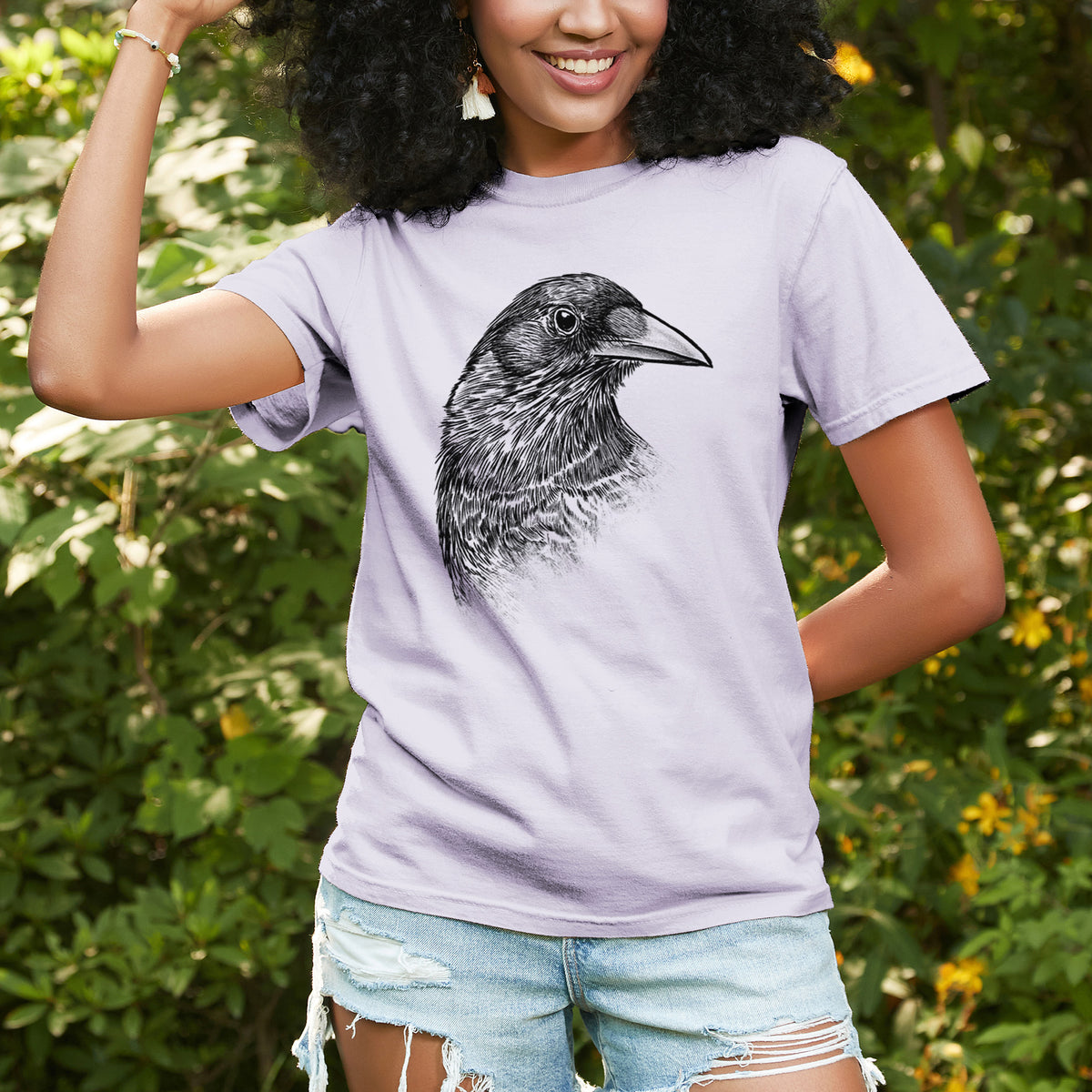 Woman wearing a Comfort Colors t-shirt printed with a detailed black ink portrait of a crow's head in profile, outdoors.