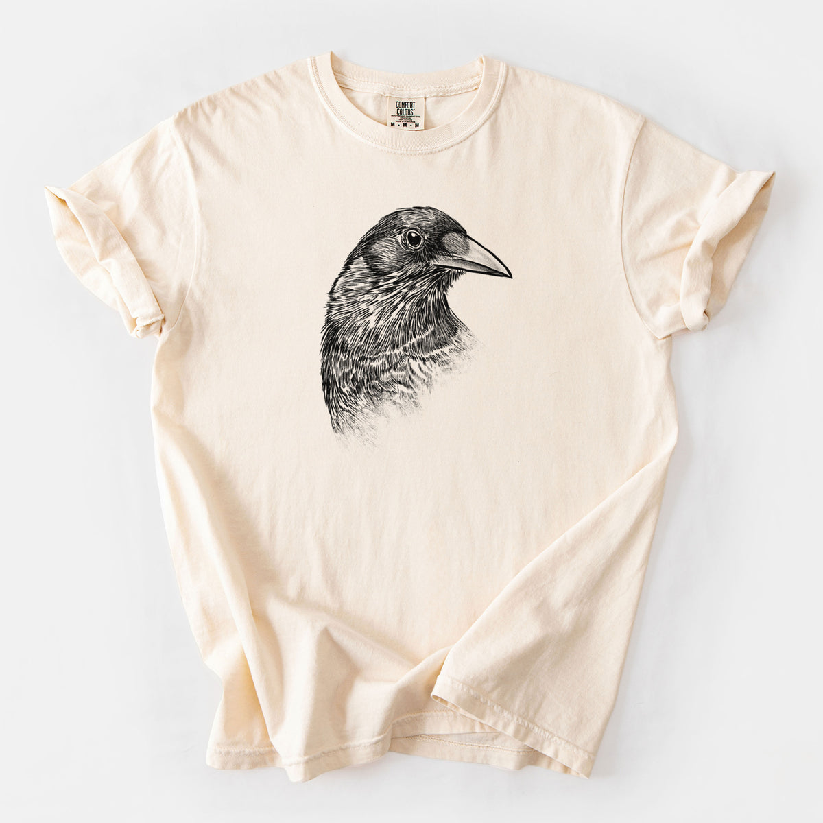Ivory Comfort Colors t-shirt laid flat, printed with a detailed black ink portrait of a crow's head in profile.