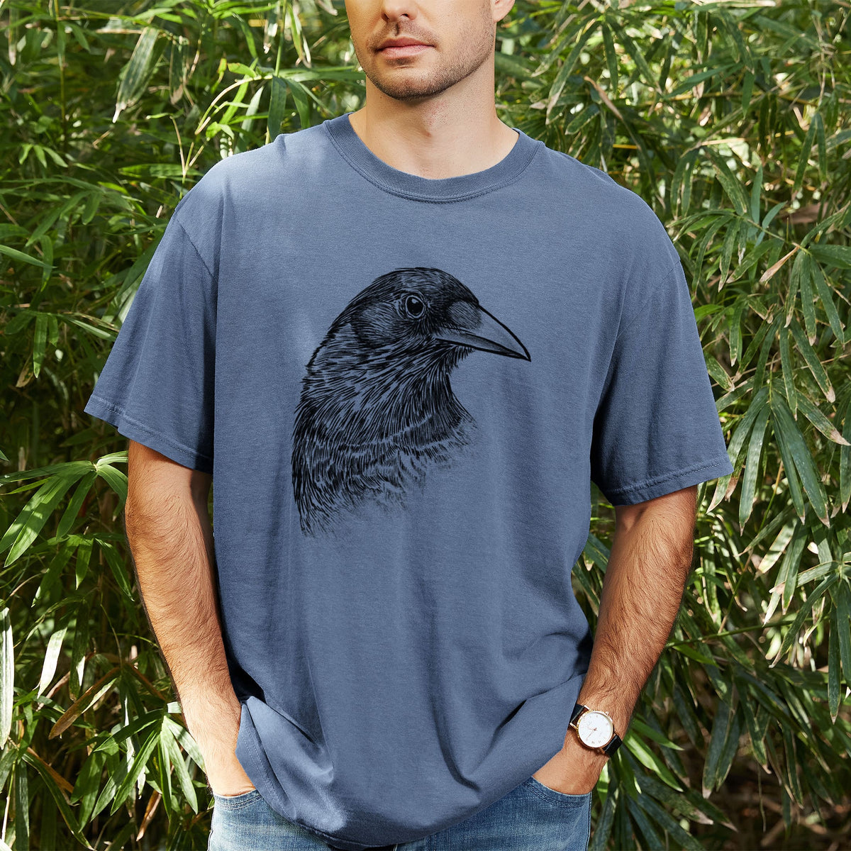 Man wearing a Comfort Colors t-shirt printed with a detailed black ink portrait of a crow's head in profile, outdoors.