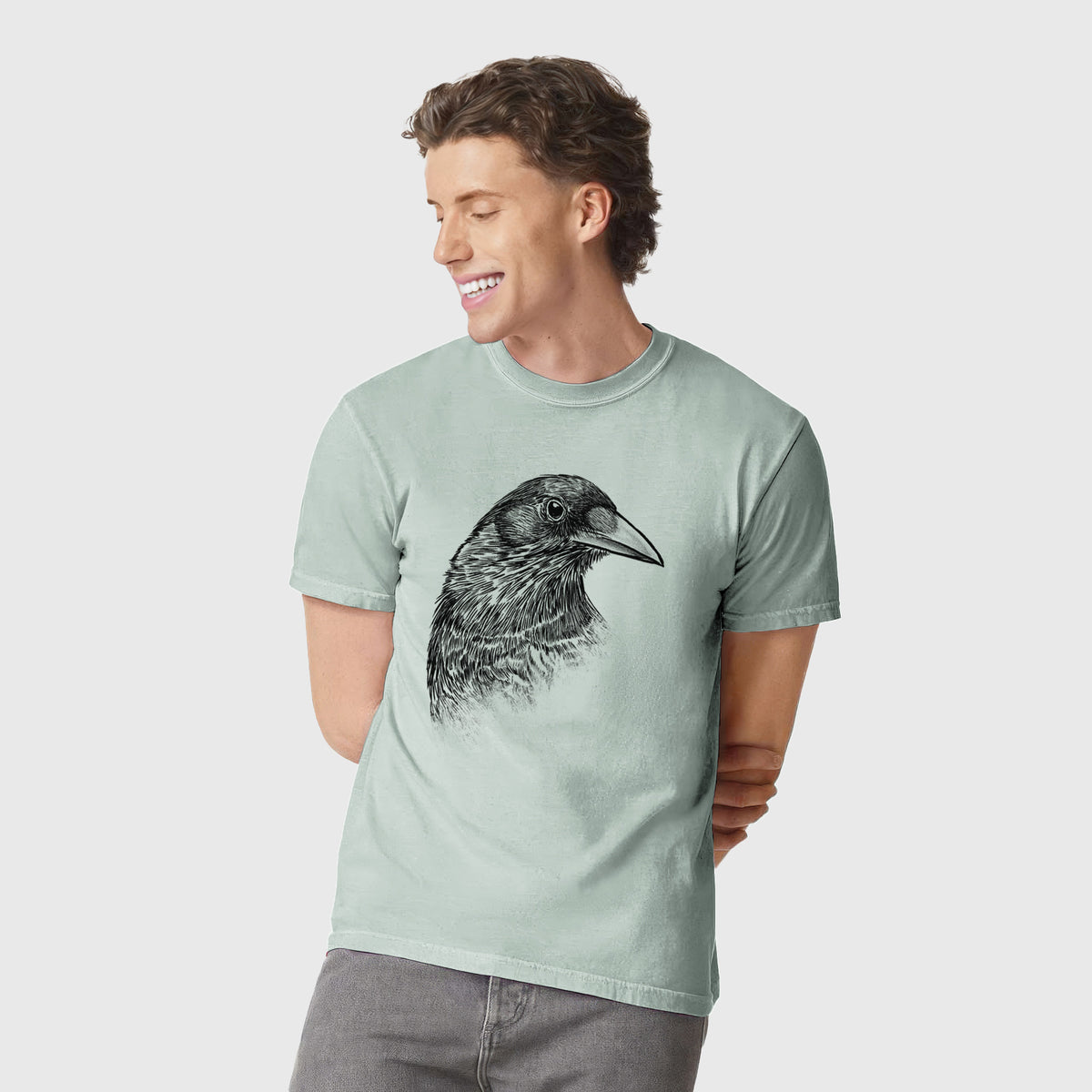 Man wearing a Comfort Colors t-shirt printed with a detailed black ink portrait of a crow's head in profile.