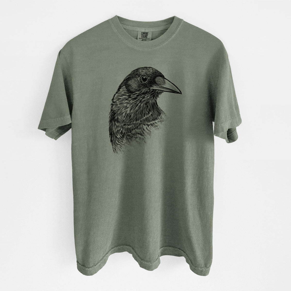 Moss green Comfort Colors t-shirt laid flat, printed with a detailed black ink portrait of a crow's head in profile.