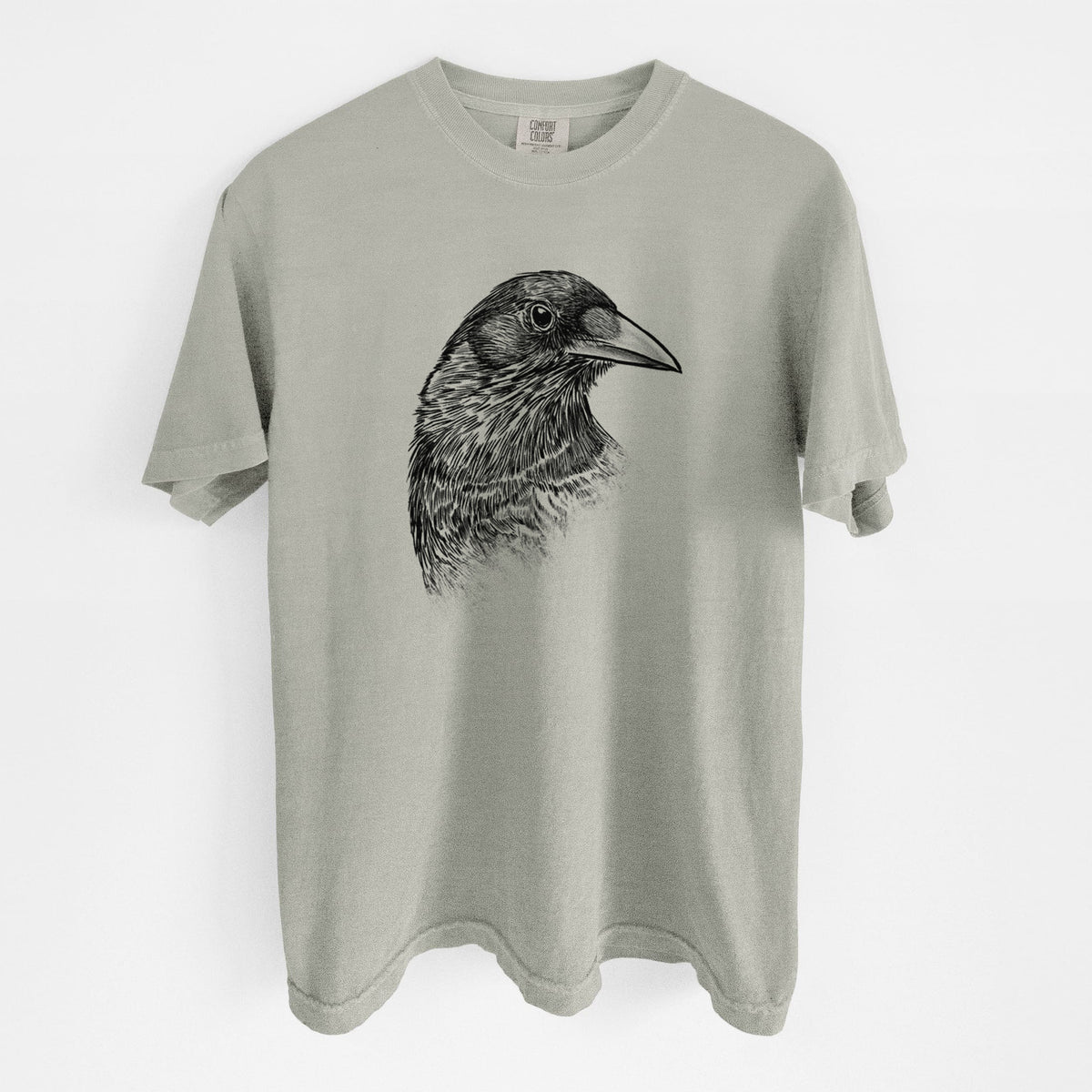 Sandstone beige Comfort Colors t-shirt laid flat, printed with a detailed black ink portrait of a crow's head in profile.