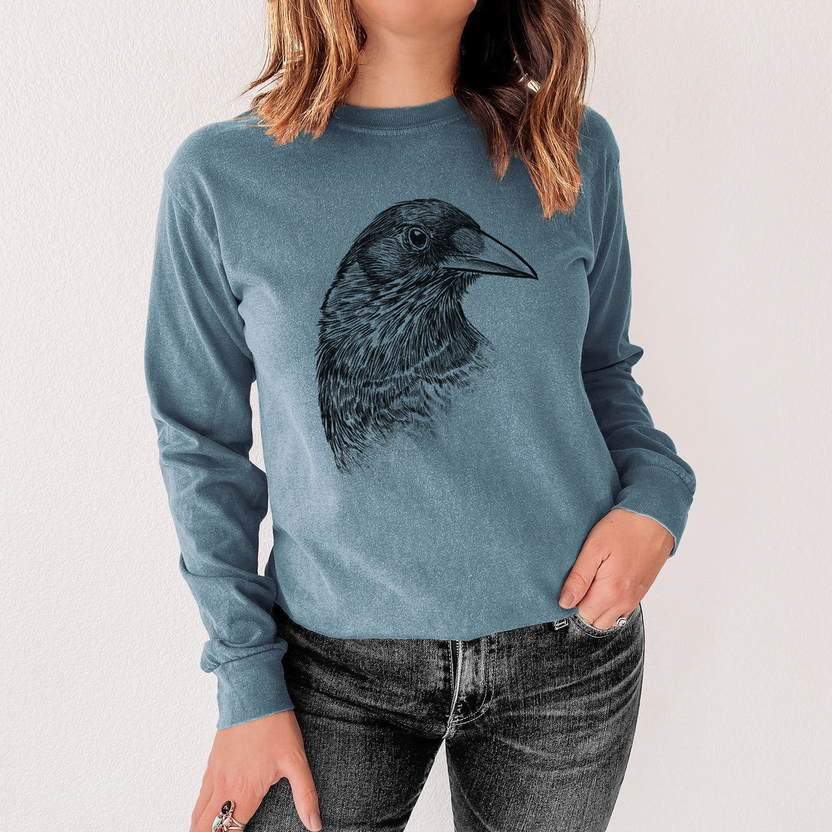 Steel blue Comfort Colors long sleeve shirt printed with a detailed black ink portrait of a crow's head in profile, shown in a lifestyle photo.