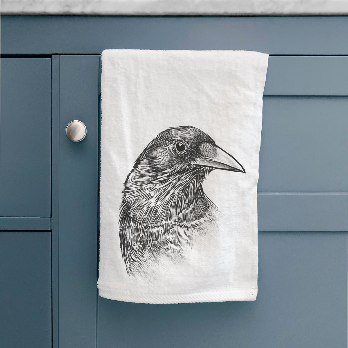 White hand towel printed with a detailed black ink portrait of a crow's head in profile, hanging on a cabinet door.