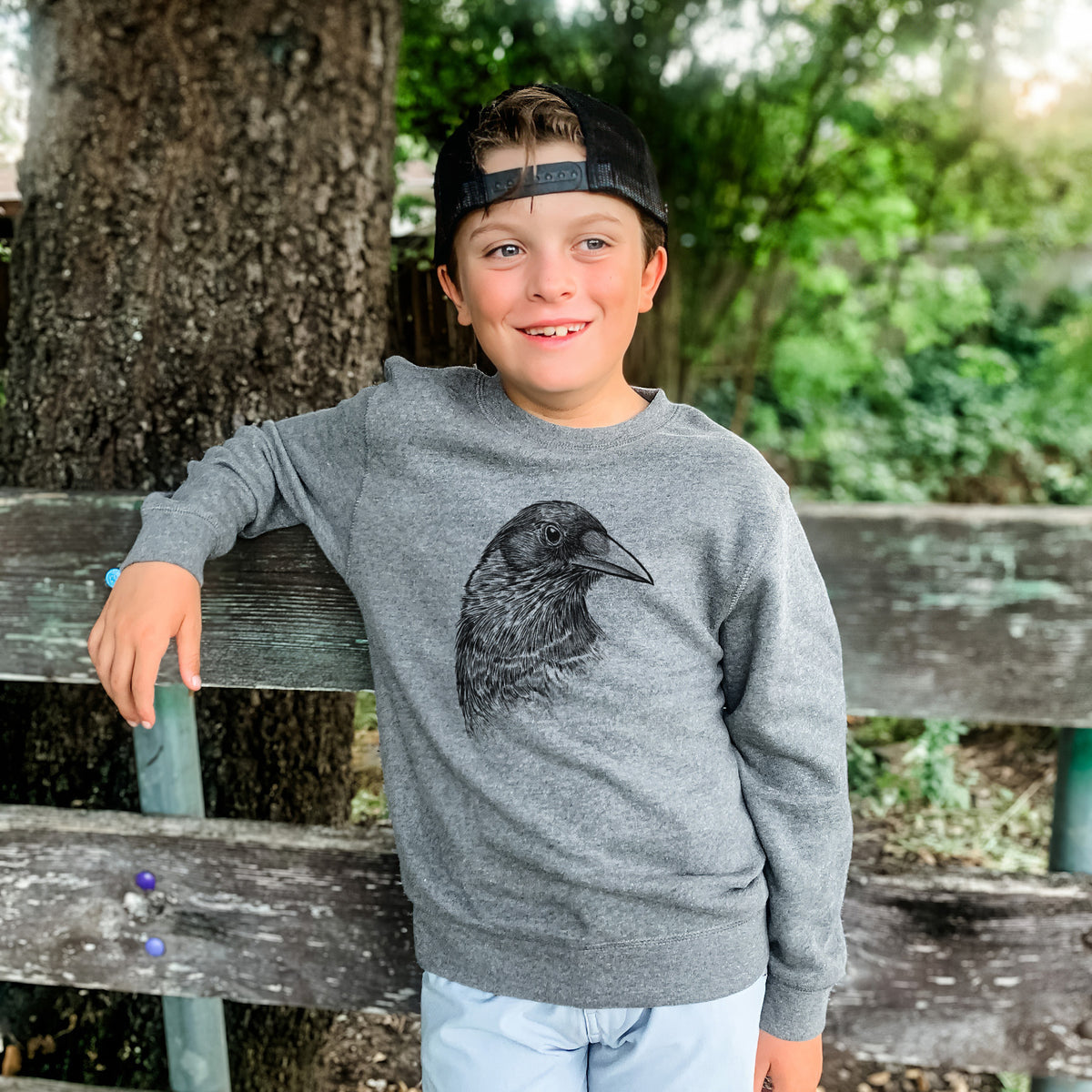 Boy wearing a youth crewneck sweatshirt printed with a detailed black ink portrait of a crow's head in profile, outdoors.