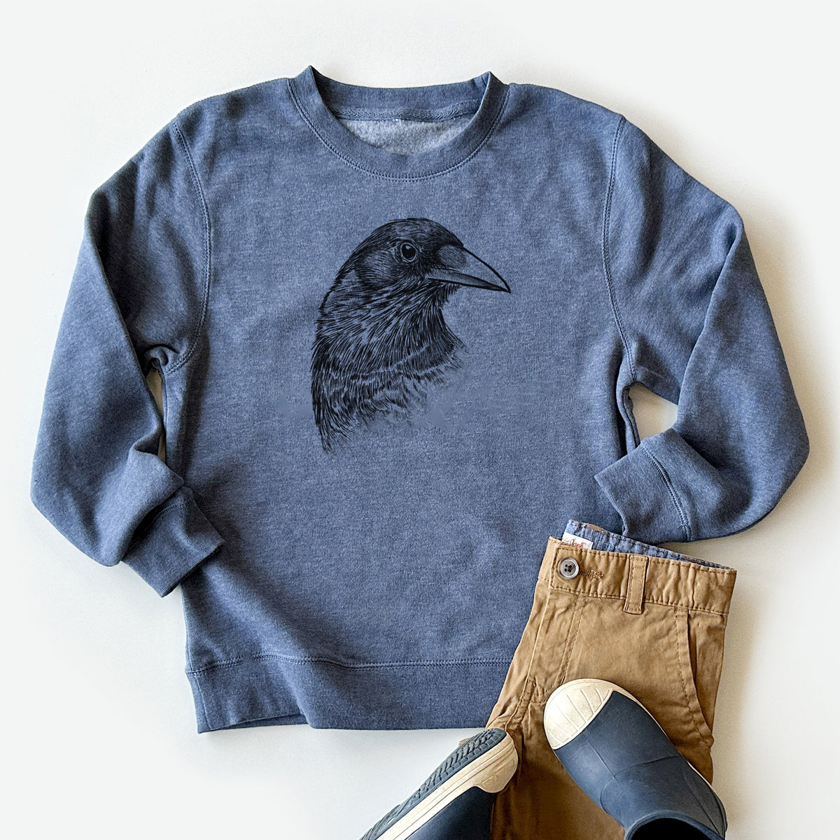 Youth crewneck sweatshirt printed with a detailed black ink portrait of a crow's head in profile, styled flat with khaki pants and shoes.