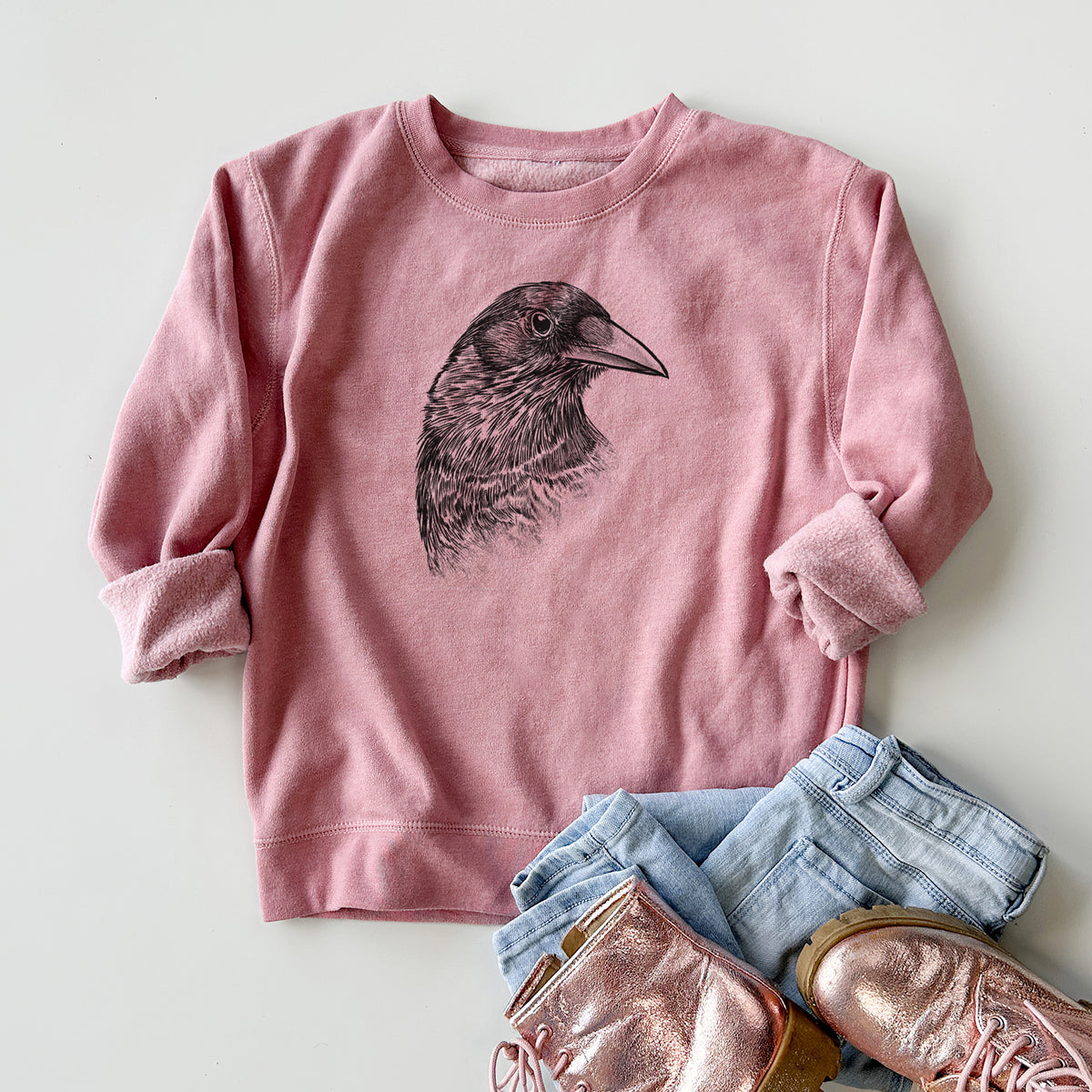Youth crewneck sweatshirt printed with a detailed black ink portrait of a crow's head in profile, styled flat with jeans and boots.