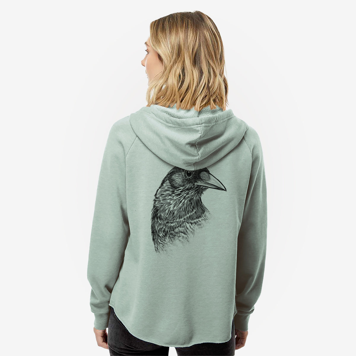Woman wearing a sage green women's Cali Wave zip-up sweatshirt printed with a detailed black ink portrait of a crow's head in profile, seen from behind.