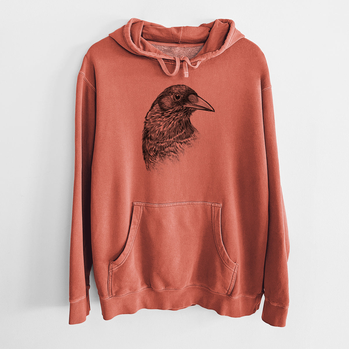 Amber orange pigment dyed hoodie laid flat, printed with a detailed black ink portrait of a crow's head in profile.