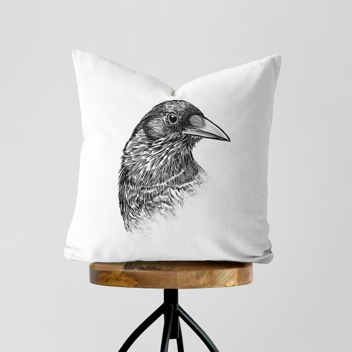 White pillow cover printed with a detailed black ink portrait of a crow's head in profile, on a wooden stool.
