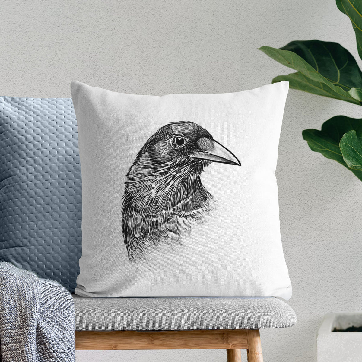 White pillow cover printed with a detailed black ink portrait of a crow's head in profile, on a bench beside plants.