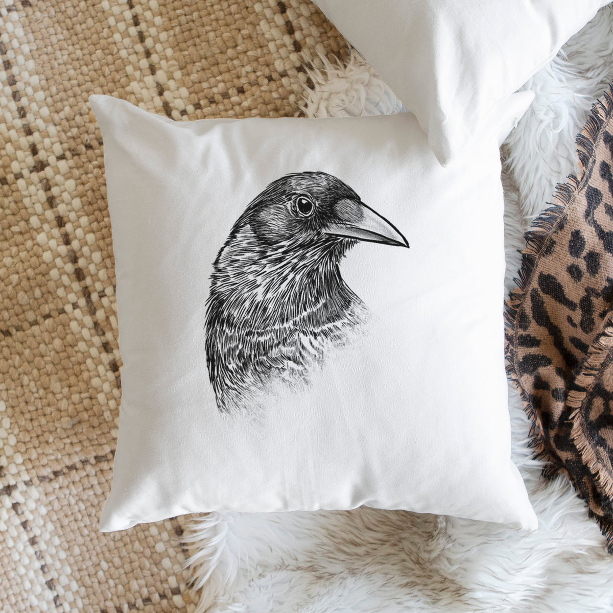 White pillow cover printed with a detailed black ink portrait of a crow's head in profile, on a woven chair.