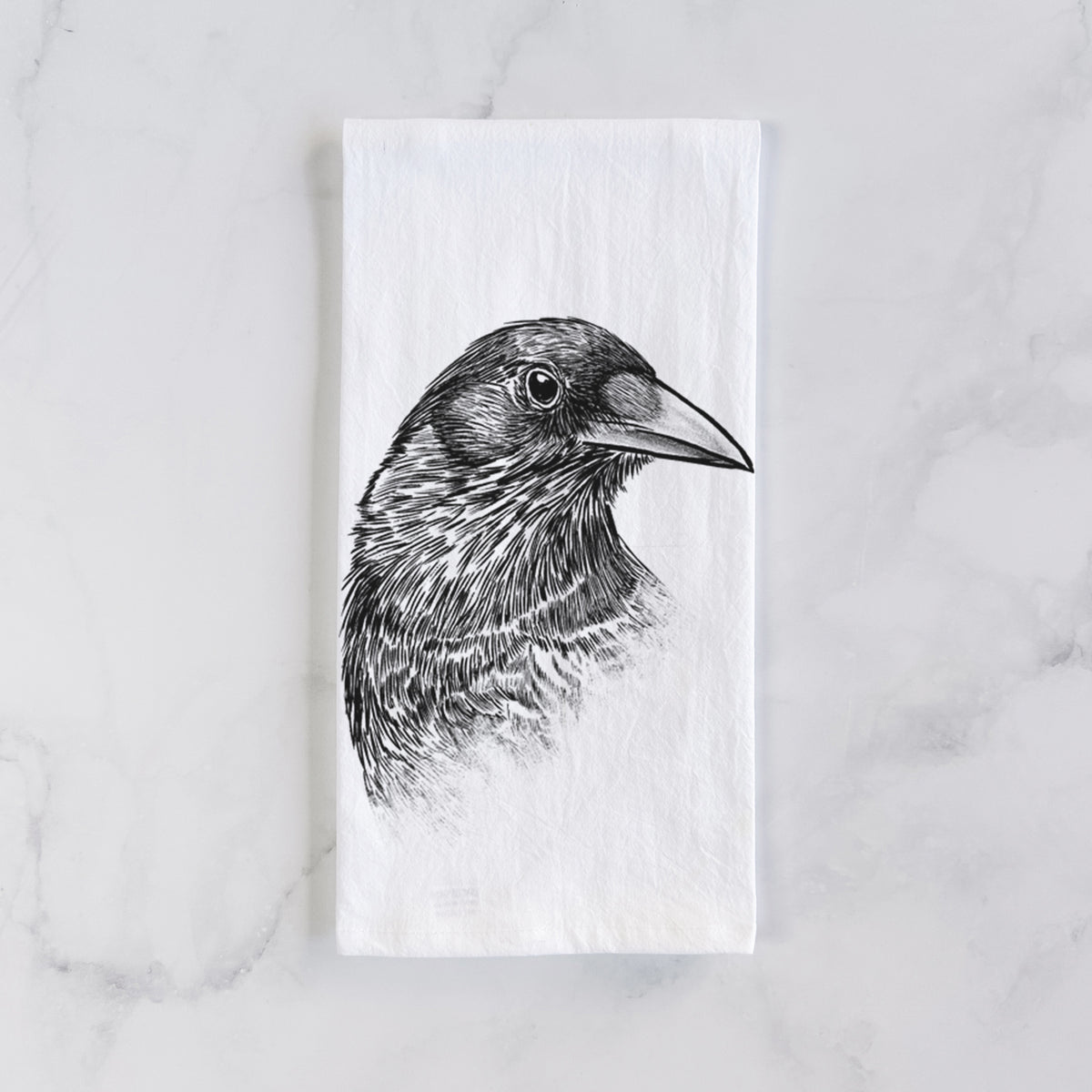 White cotton tea towel laid flat on marble, printed with a detailed black ink portrait of a crow's head in profile.