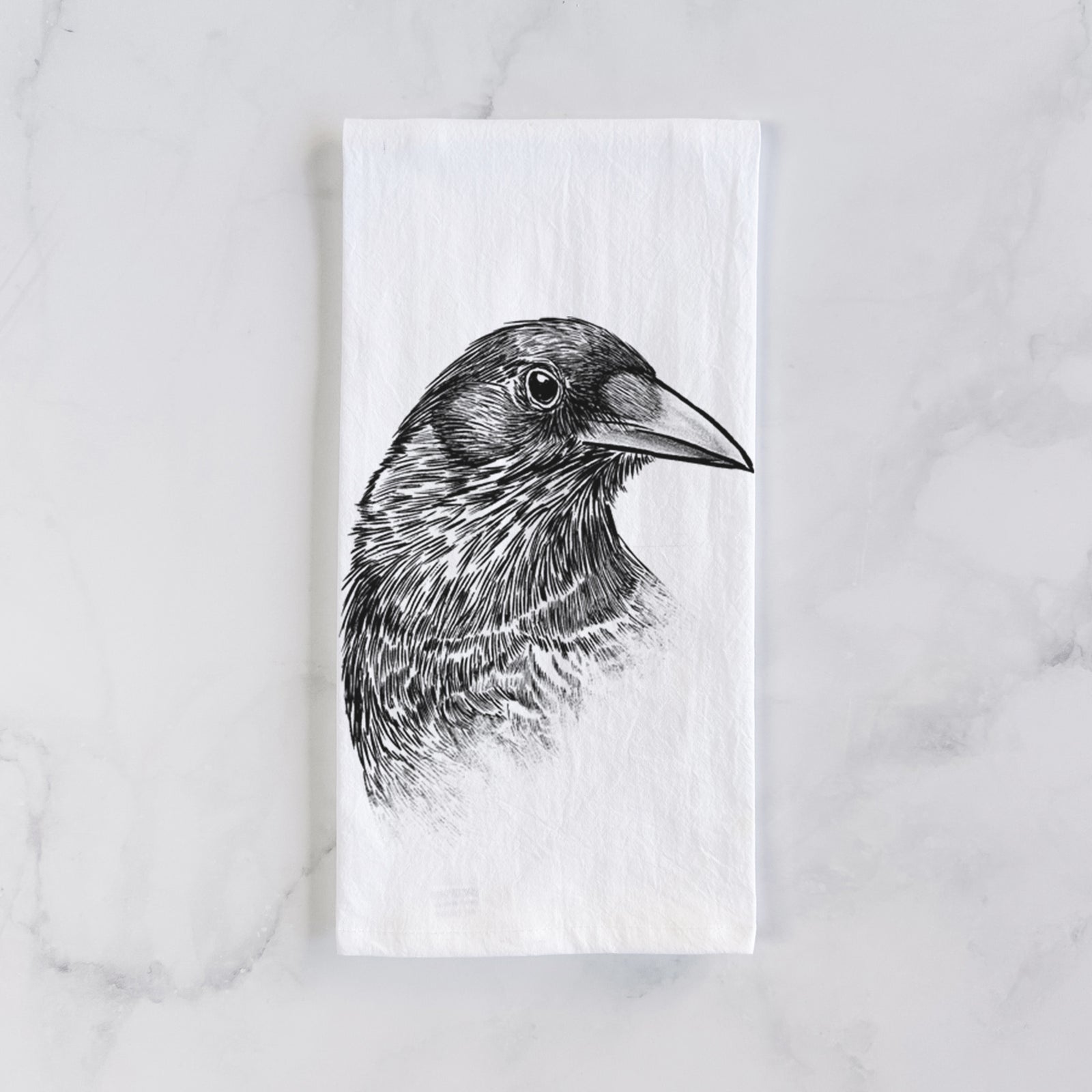 White cotton tea towel printed with a detailed black ink portrait of a crow's head in profile, on a marble counter beside a round wooden cutting board and greenery.