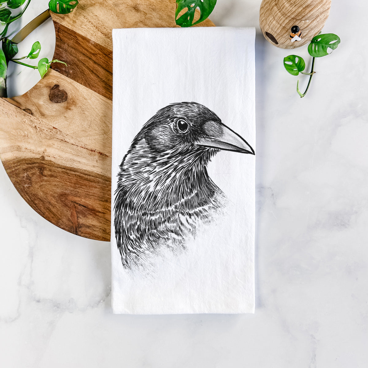 White cotton tea towel printed with a detailed black ink portrait of a crow's head in profile, on a marble counter beside a round wooden cutting board and greenery.