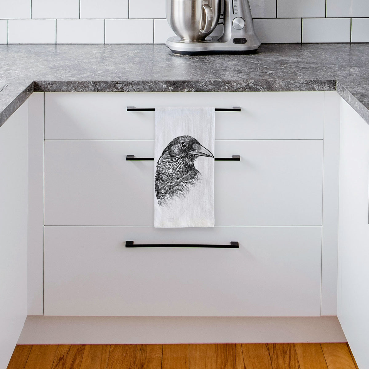 White tea towel printed with a detailed black ink portrait of a crow's head in profile, hanging from a drawer handle in a bright white kitchen.