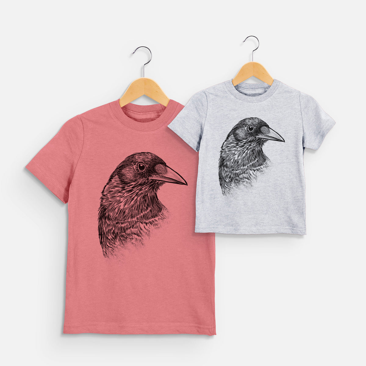 Two youth t-shirts in different colors on hangers, printed with a detailed black ink portrait of a crow's head in profile.