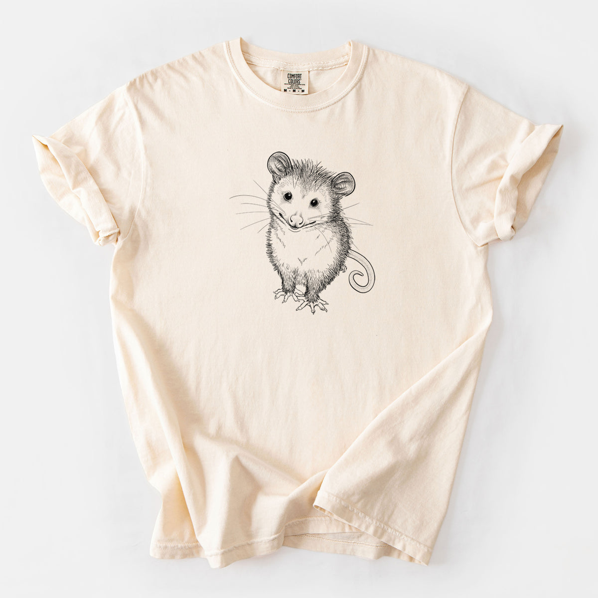 Ivory Comfort Colors t-shirt laid flat, printed with a black ink illustration of a wide-eyed baby opossum sitting.
