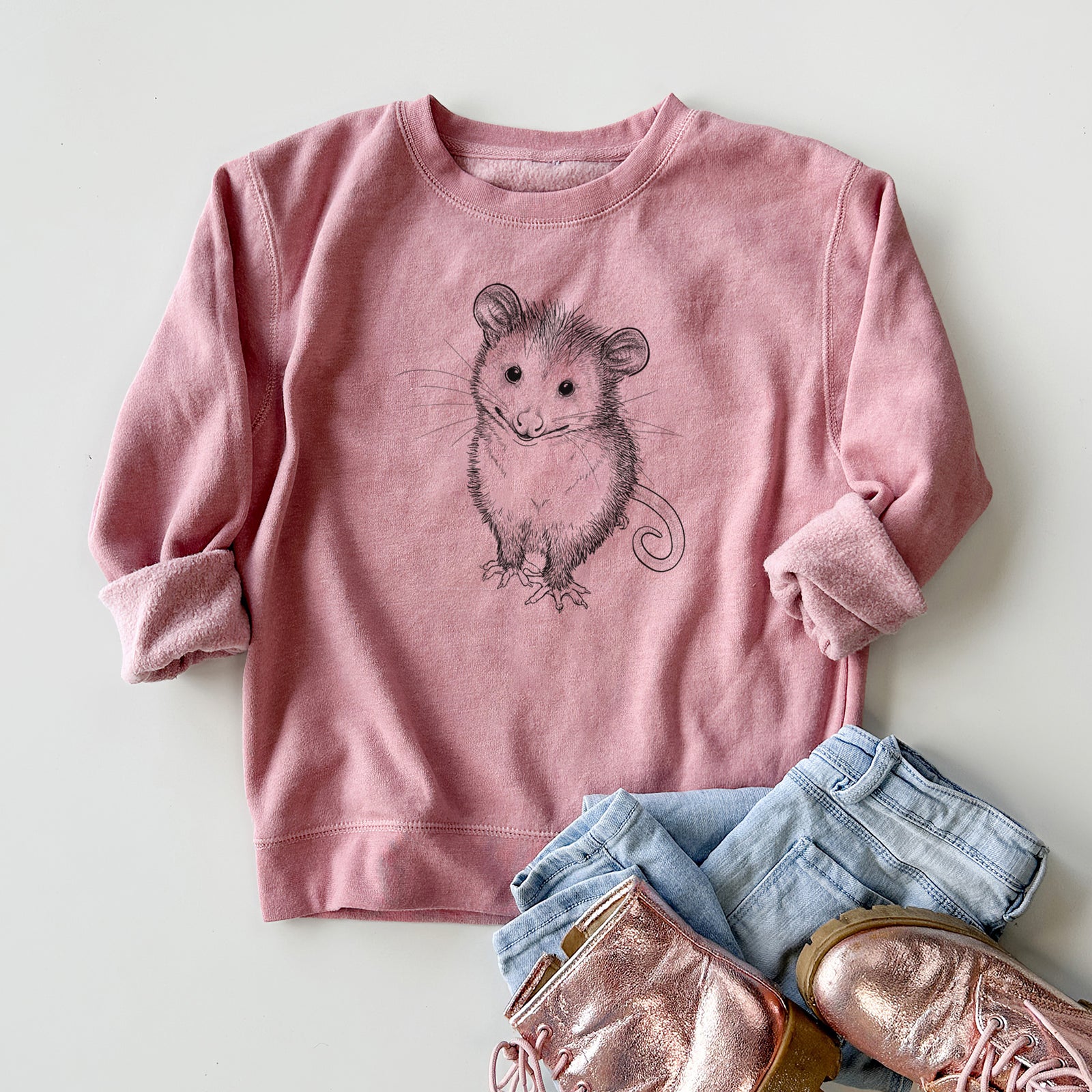 Boy wearing a youth crewneck sweatshirt printed with a black ink illustration of a wide-eyed baby opossum sitting, outdoors.