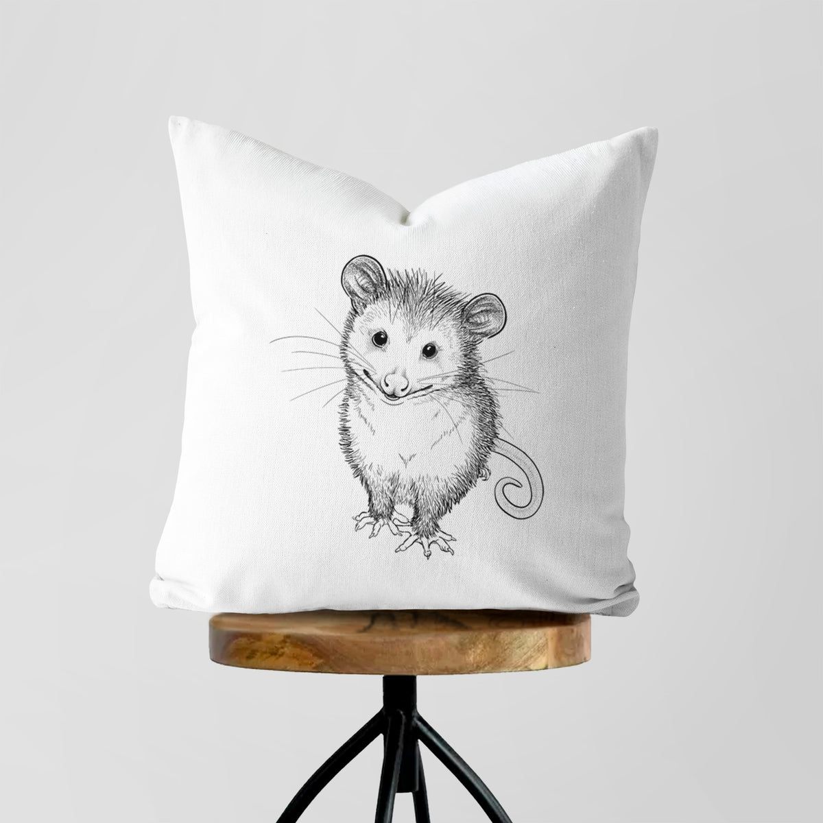 White pillow cover printed with a black ink illustration of a wide-eyed baby opossum sitting, on a wooden stool.