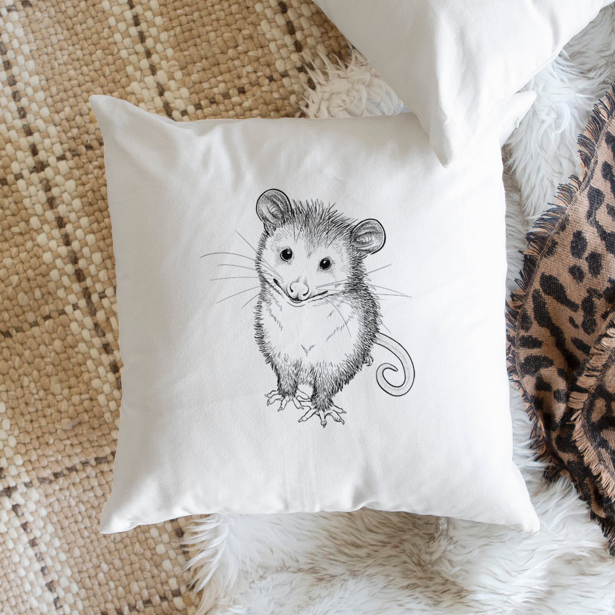 White pillow cover printed with a black ink illustration of a wide-eyed baby opossum sitting, on a woven chair.
